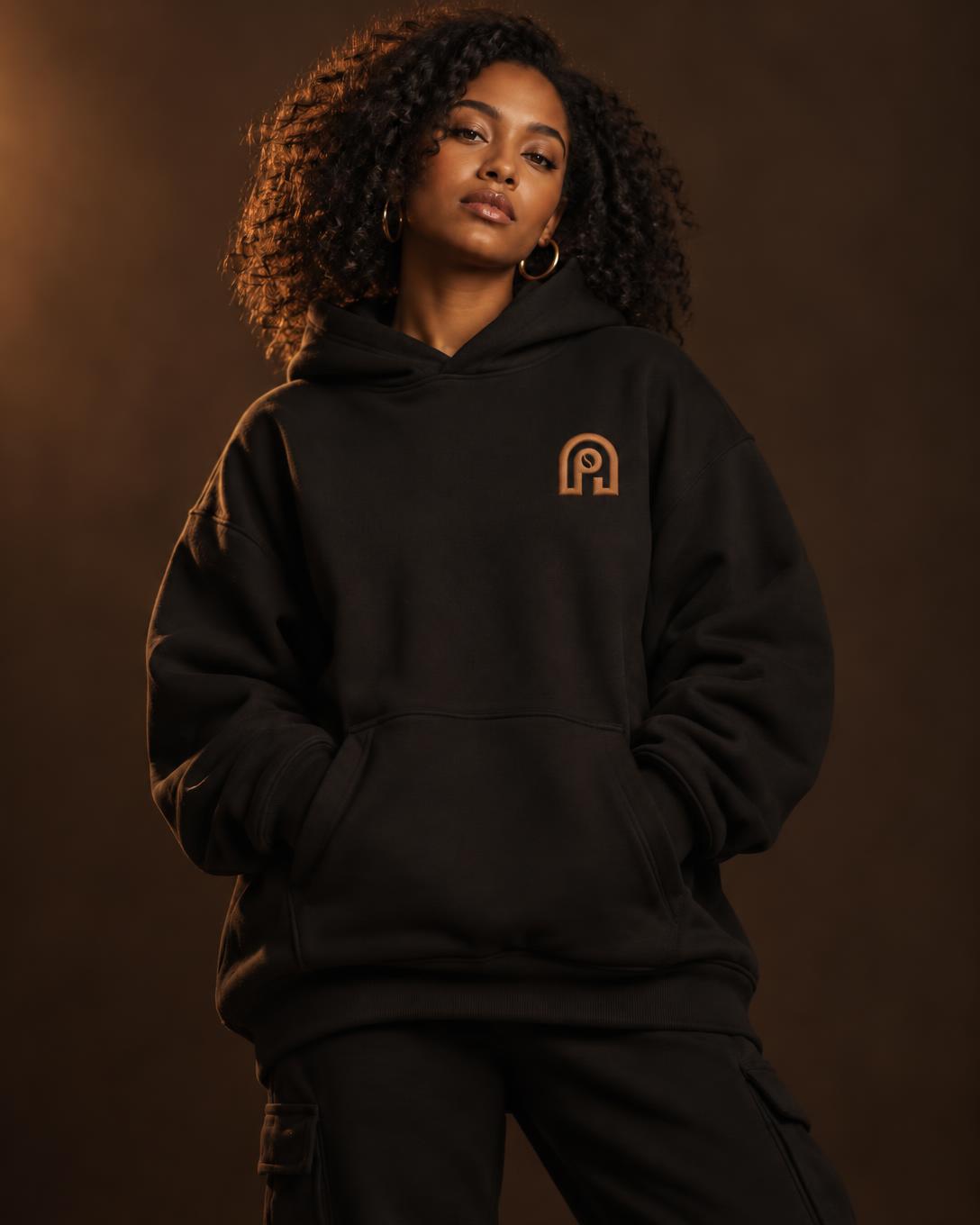 A woman with natural curls wearing an oversized long-black NAPPUCCI hoodie with the NAPPUCCI arch logo embroidered in caramel on the chest, in a warm brown studio